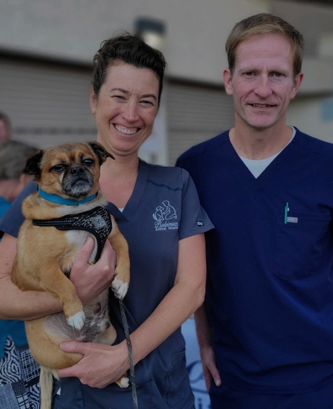 Two veterinarians, one holding a small dog, smiling outdoors.