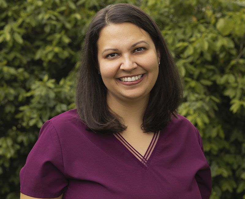 Smiling woman in a purple top standing before green foliage.
